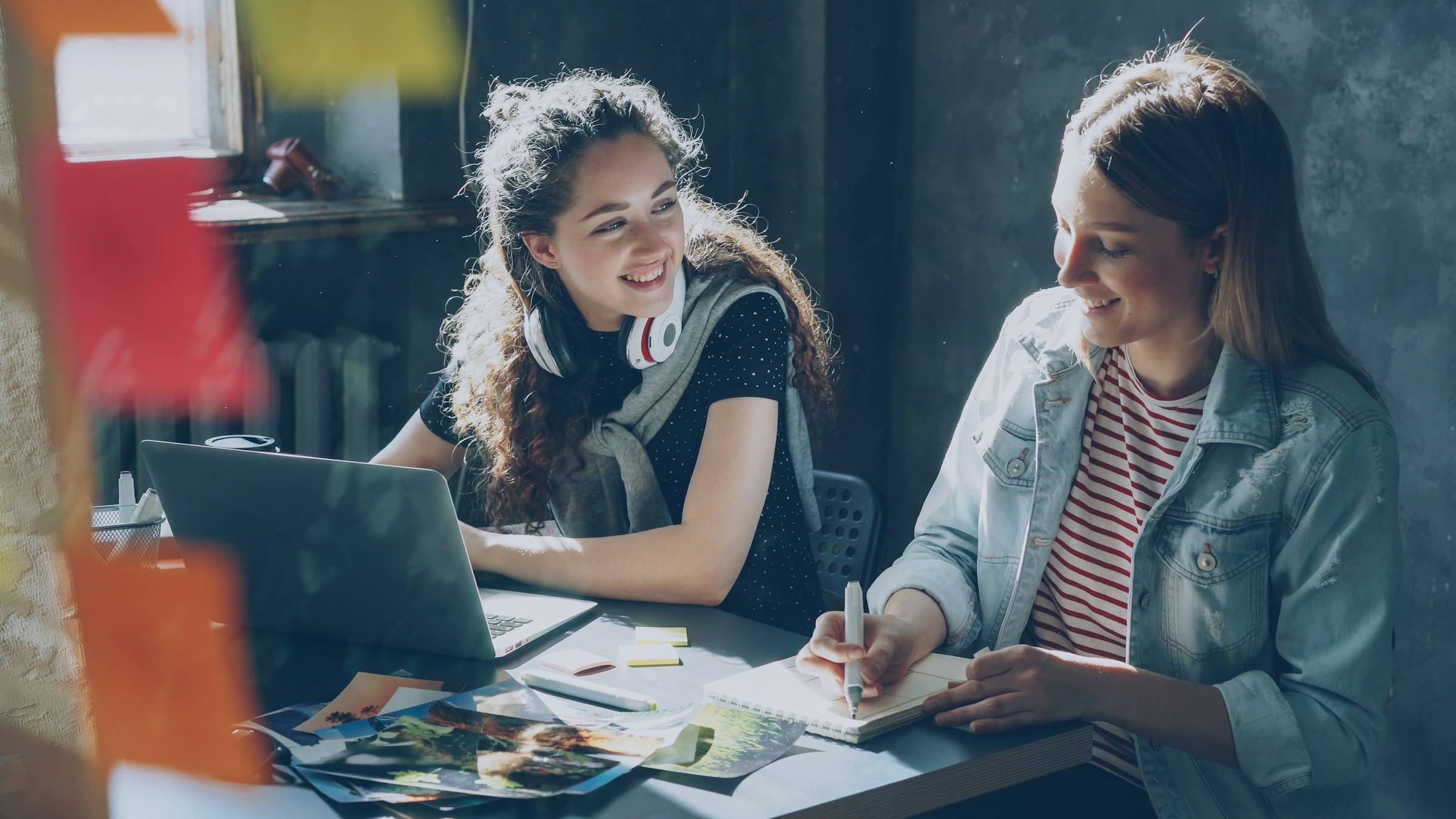 Two young women at a table, one writing in a notebook while the other smiles